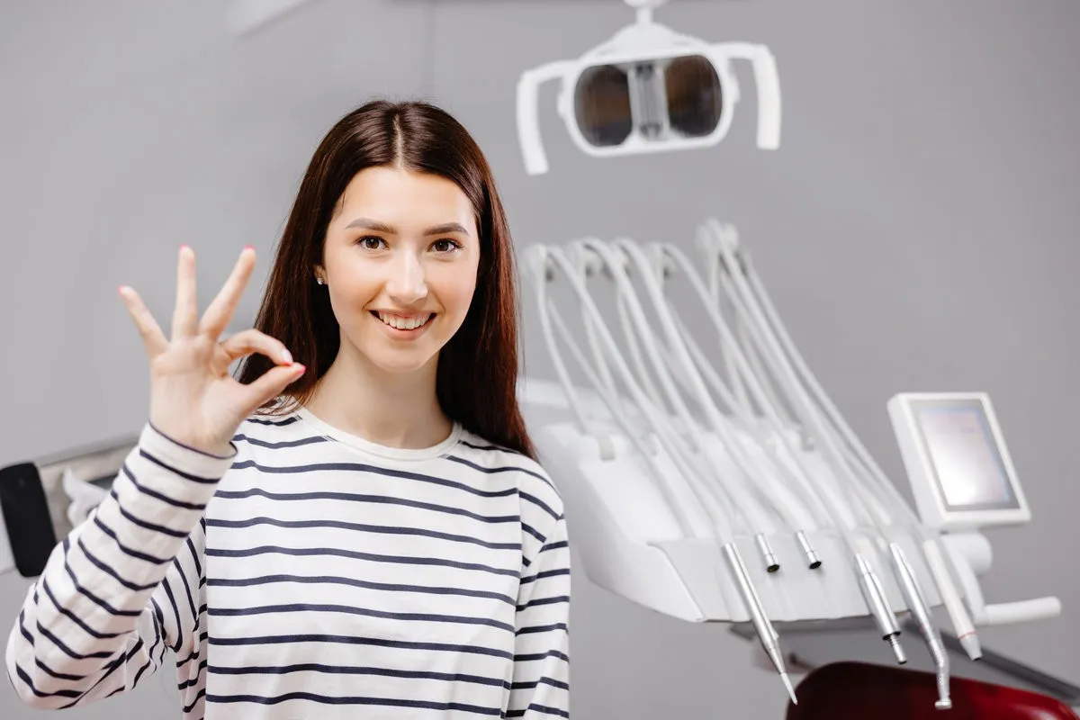 Young smiling happy girl showing okay gesture sit at dentist office chair after dental procedure. Healthcare teeth treatment.