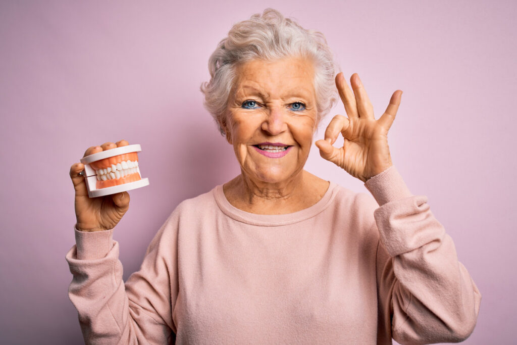 Senior beautiful grey-haired woman holding plastic denture teeth over pink background doing ok sign with fingers, excellent symbol