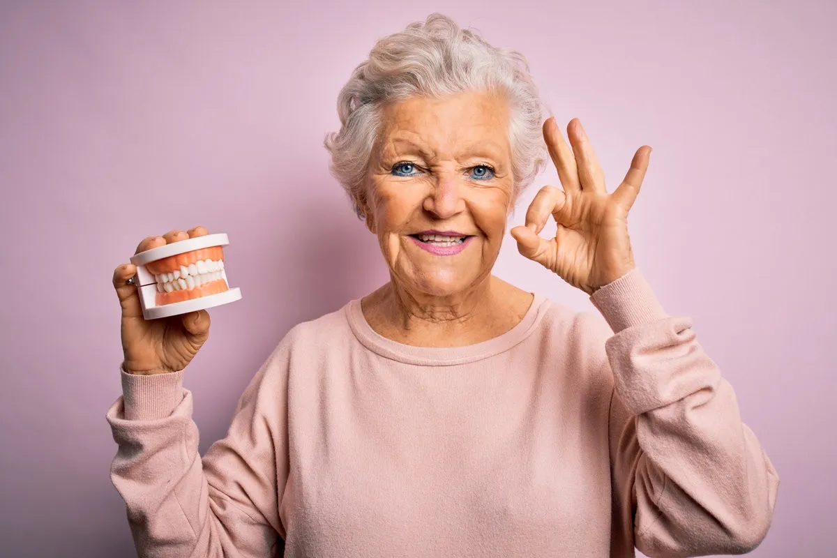 Senior beautiful grey-haired woman holding plastic denture teeth over pink background doing ok sign with fingers, excellent symbol