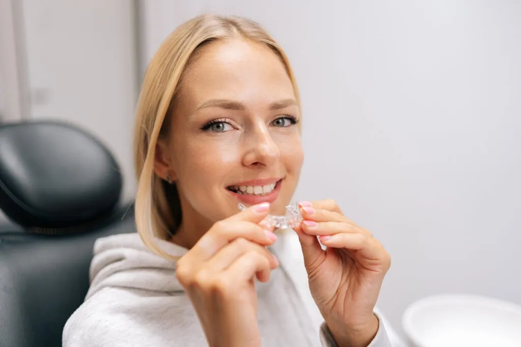 Closeup portrait of smiling blonde female patient holding invisible braces aligner sitting on chair in dentistry clinic. Close up of mobile orthodontic appliance for dental correction.